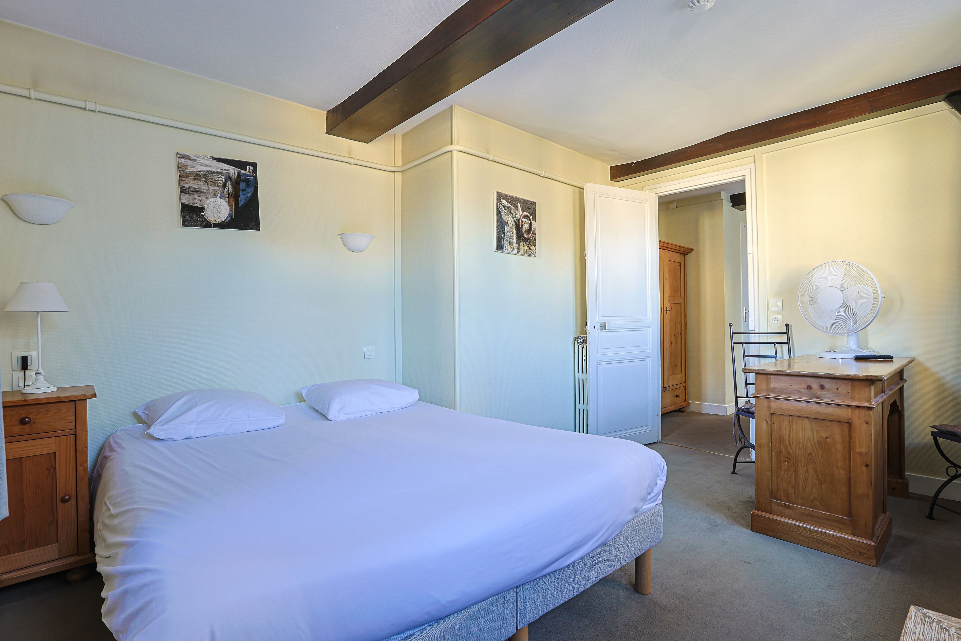 Un dormitorio con cama, una mesita de noche de madera, un escritorio con silla y una puerta que da a otra habitación.