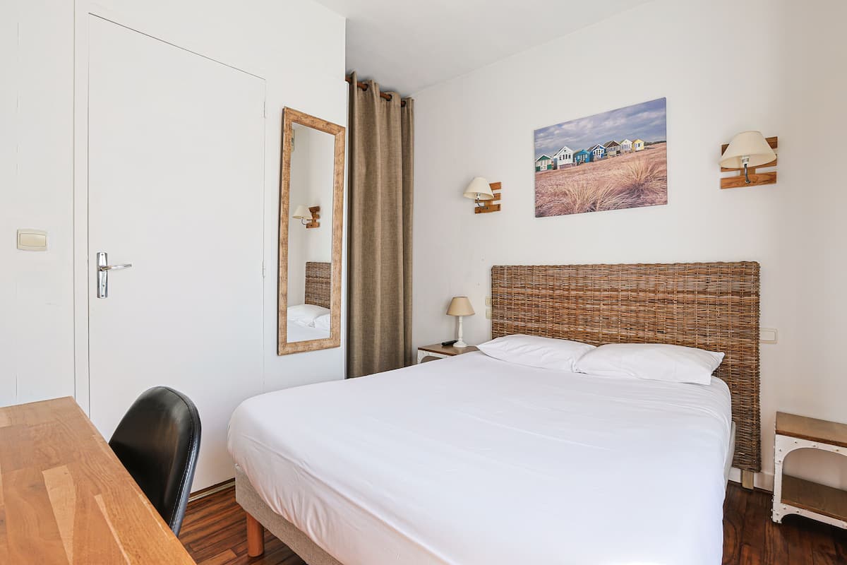 A hotel room with a large bed, a wooden desk, a chair, a mirror, a picture on the wall, and a lamp.