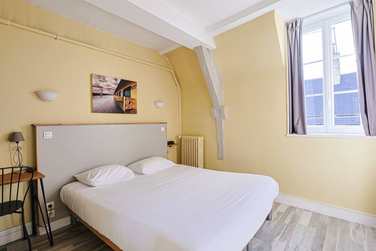 A bedroom with a large bed, a bedside table, a chair, a radiator, a window with curtains, and a wall-mounted picture.