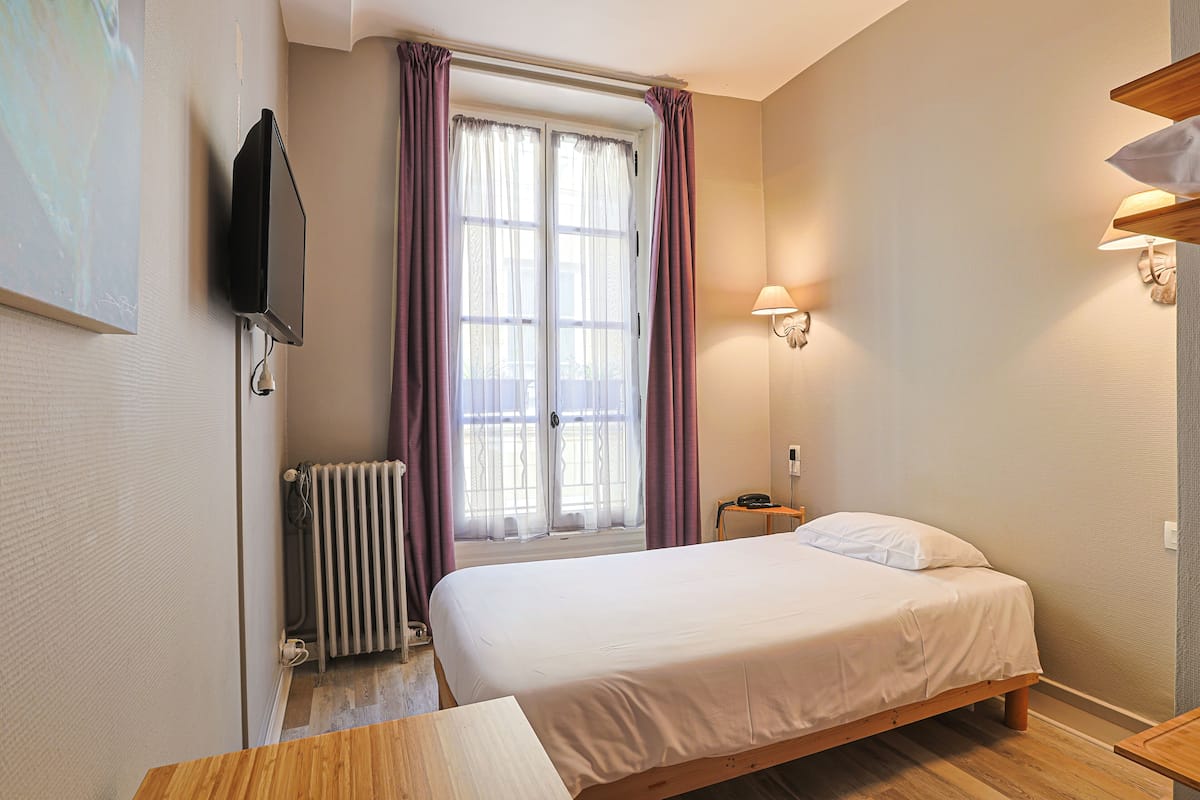 A bedroom with a bed, a radiator, a TV mounted on the wall, and a window with curtains.