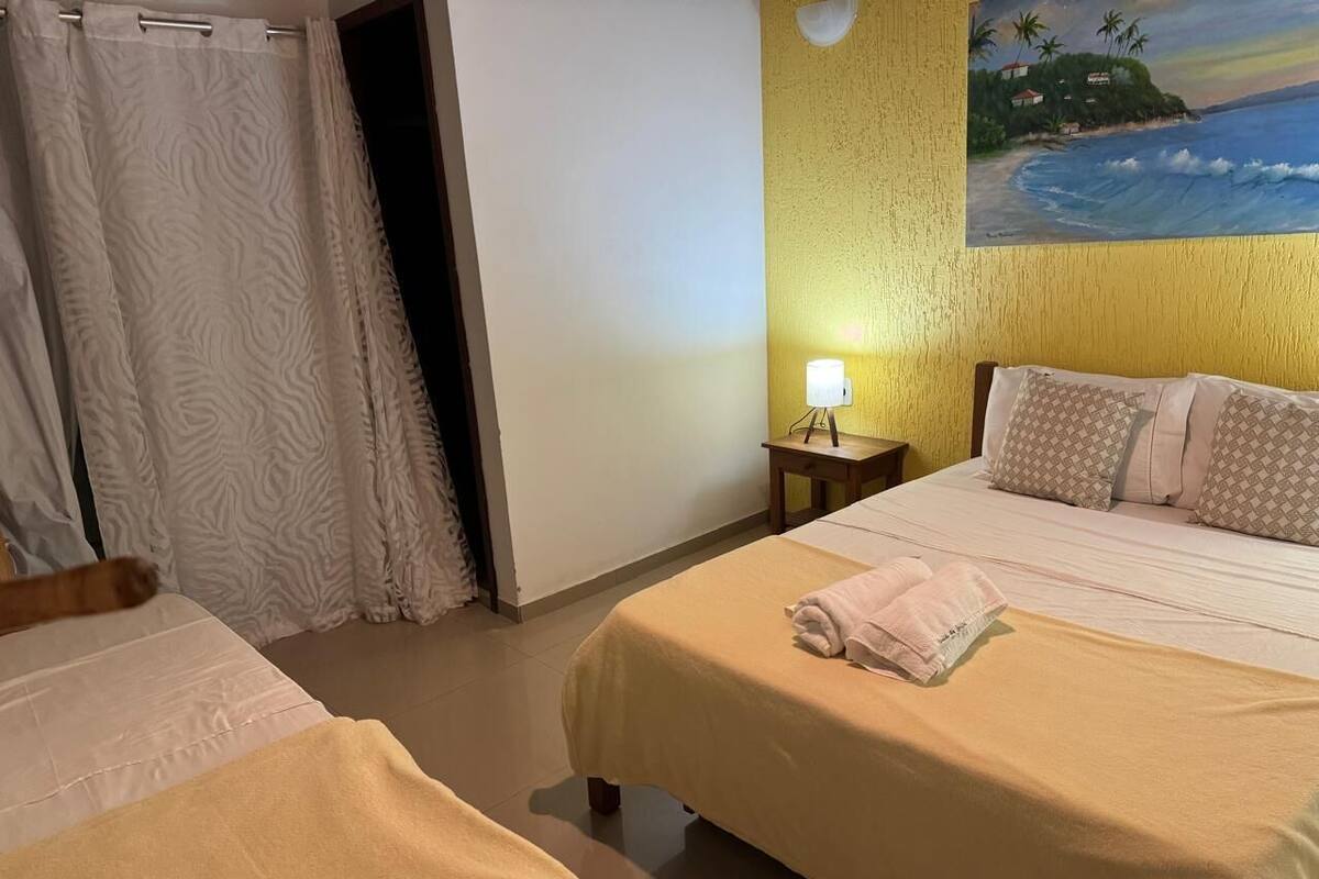 Habitación de hotel con dos camas, un cuadro de una playa, una mesita de noche con lámpara y una cortina.