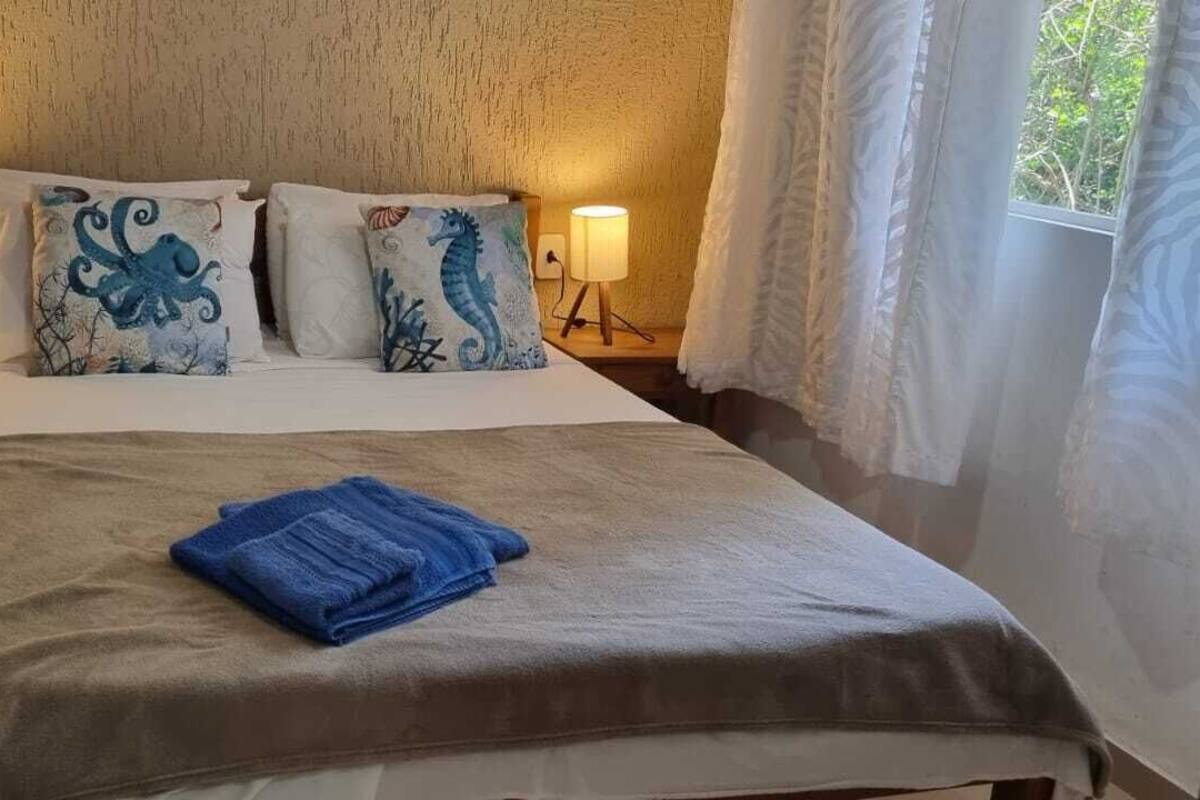 Una cama bien tendida con una toalla azul, dos almohadones decorativos con motivos marinos, dos lámparas de noche y una ventana con cortinas.
