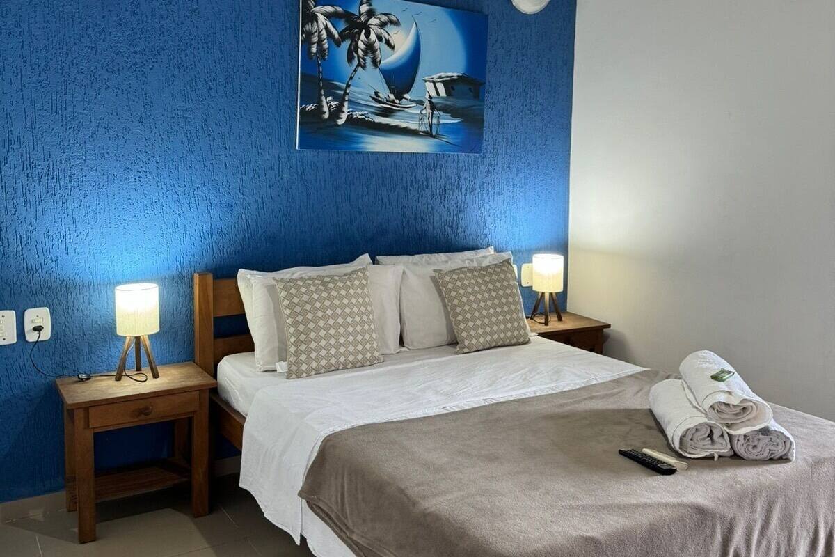 Un dormitorio con pared azul con textura, un cuadro de un velero, una cama con ropa blanca, dos lámparas de noche y un ventilador de techo.