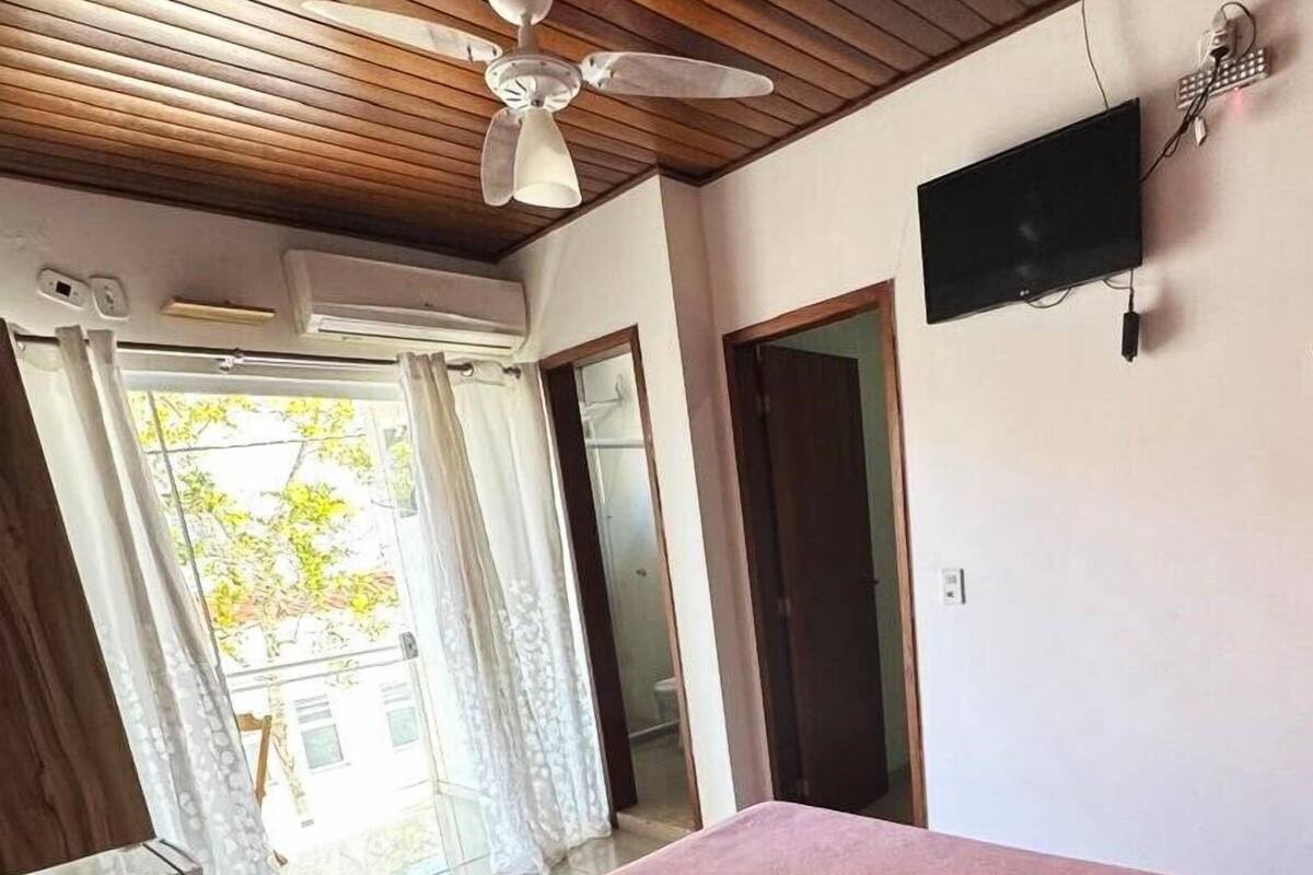 Un dormitorio con techo de madera, una cama con colcha rosa, un televisor en la pared y una ventana con cortinas.