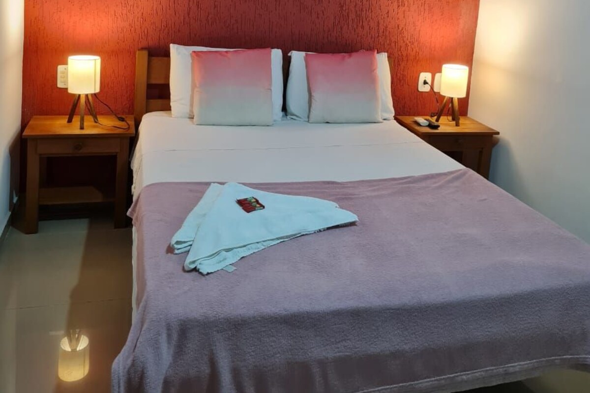 A bedroom with a wooden ceiling, a bed with a pink blanket, two bedside tables with lamps, and a floral painting on the wall.