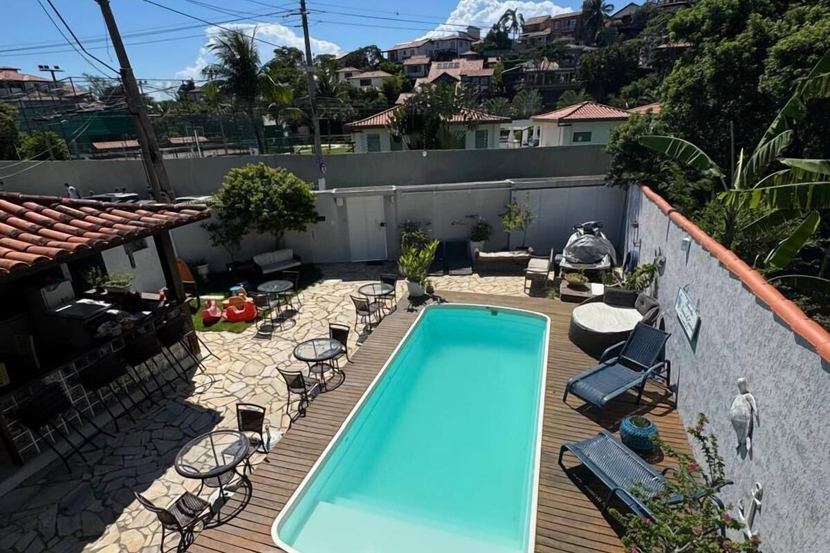 Una piscina rectangular con muelle de madera y sillas de descanso, rodeada por un muro blanco y plantas en macetas.