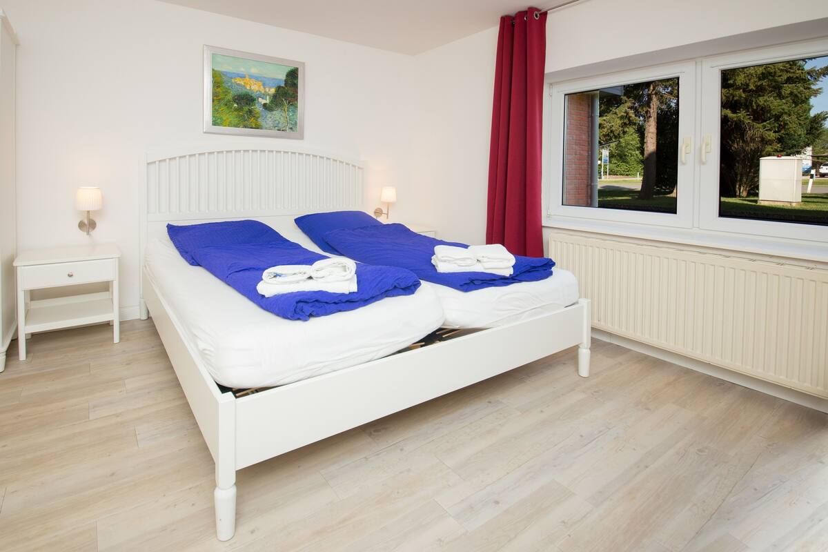 Una cama doble con ropa de cama blanca y almohadas azules, una mesita de noche con lámpara, una ventana con cortinas rojas y un cuadro en la pared.