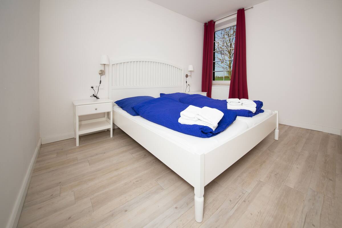 Un dormitorio con cama blanca, ropa de cama azul, una mesita de noche blanca, una cortina roja y una ventana con vistas.