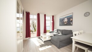 A living room with a gray sofa, white coffee table, and dining table. There are red curtains on the windows, and a clock on the wall.