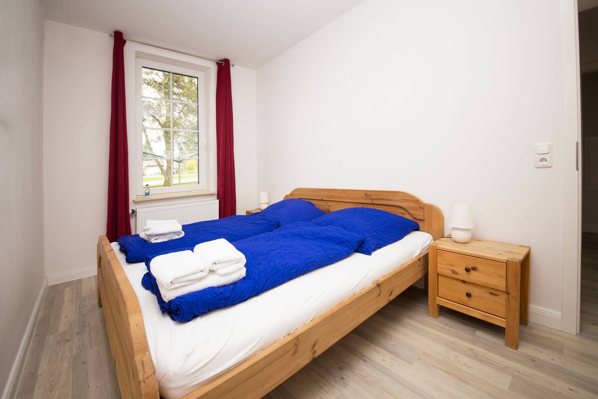 Una cama individual con ropa de cama azul, una mesita de noche de madera con una lámpara y una ventana con cortinas rojas.