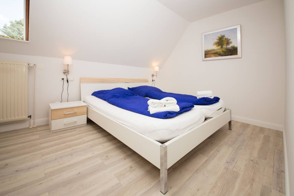Un dormitorio con cama blanca, ropa de cama azul, dos lámparas de noche, una mesita de noche y un cuadro en la pared.
