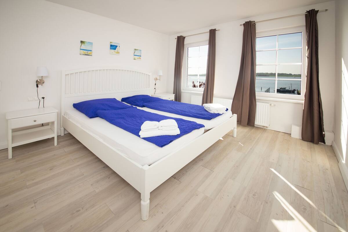 Una cama doble con ropa de cama azul, una mesita de noche, una ventana con cortinas y una obra de arte en la pared.