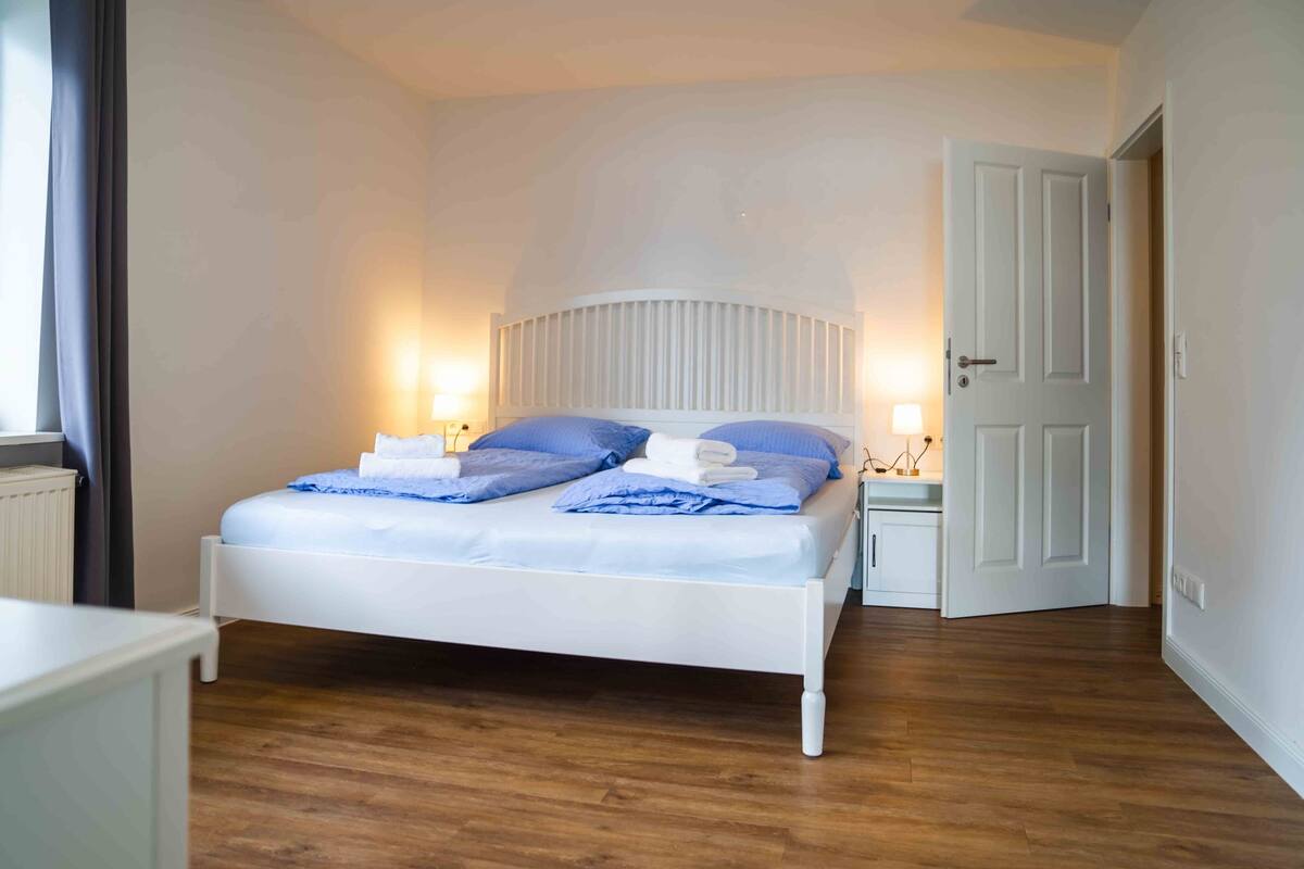 Una cama bien hecha con ropa de cama azul y blanca, un cabecero blanco y lámparas de noche.