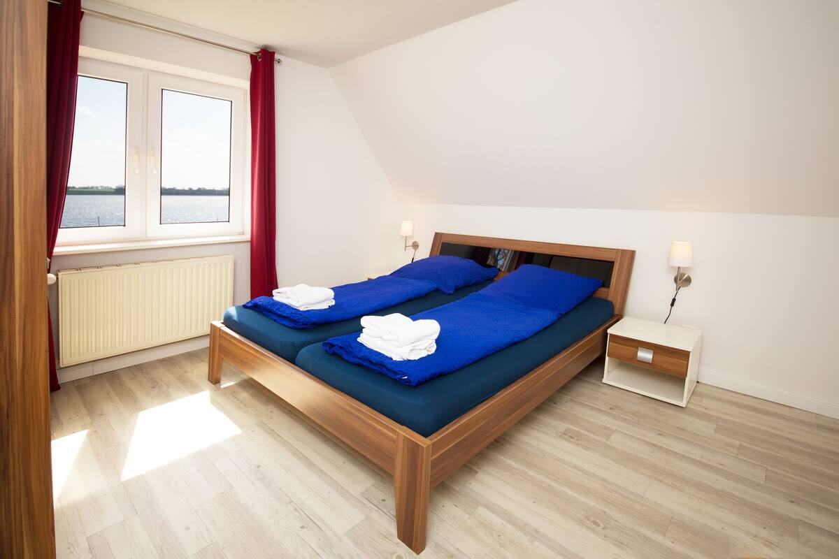 Un dormitorio con cama de madera, dos mesitas de noche, una ventana con cortinas y vista al agua.