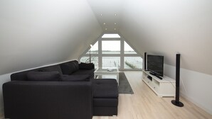 A modern living room with a large black sectional sofa, a flat-screen TV on a white stand, and a view of the water through large windows.
