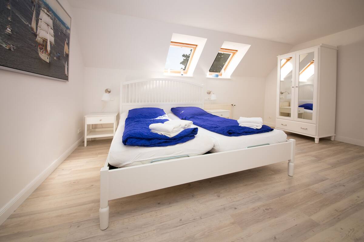 Un dormitorio con cama blanca, dos tragaluces, un armario blanco y un cuadro en la pared.