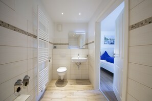 Une salle de bain moderne équipée de toilettes, d’un lavabo et d’une douche. Une fenêtre offre une vue sur les bateaux, et un balcon est agrémenté de coussins bleus.