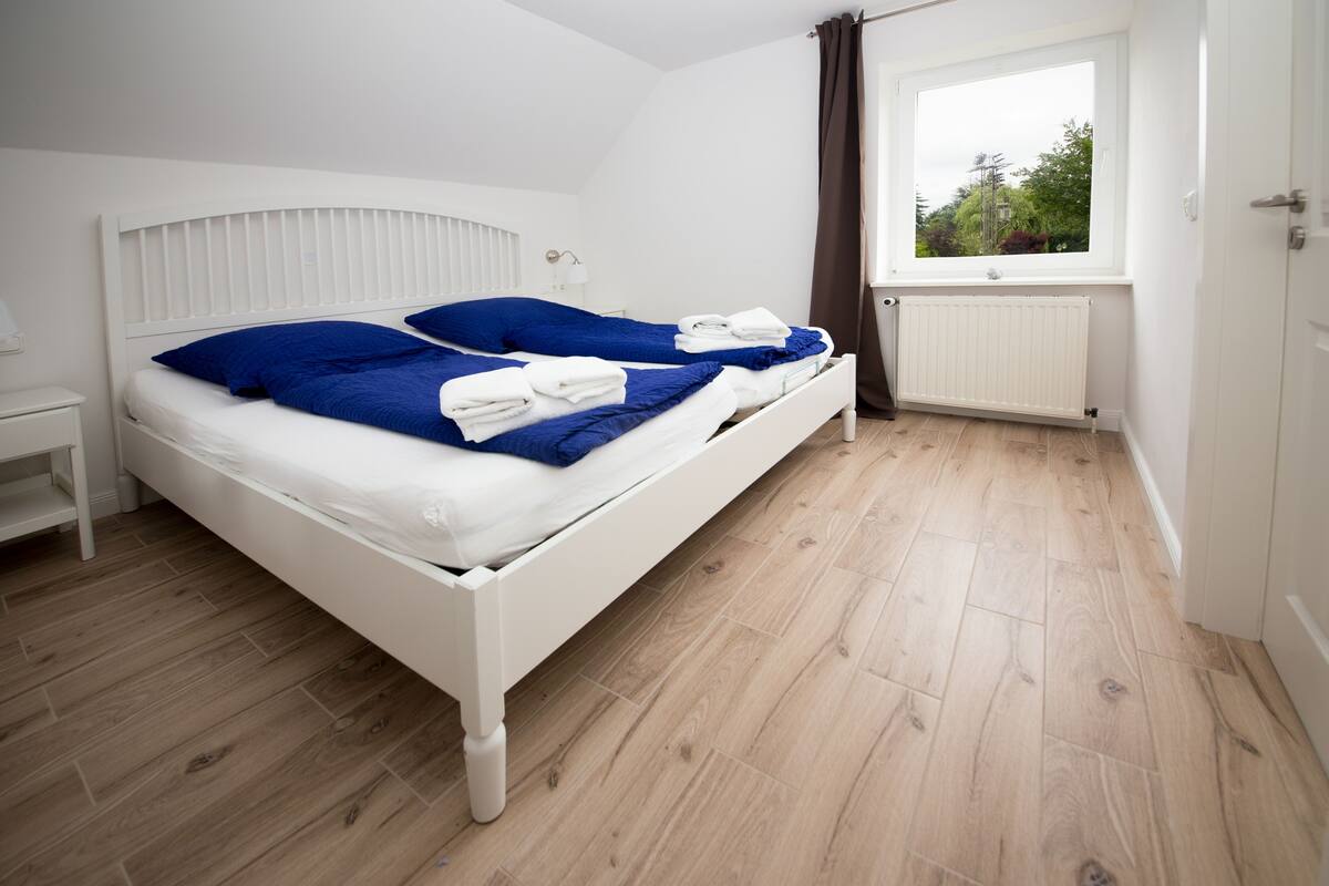 Una cama blanca con ropa de cama azul y toallas blancas, una mesita de noche blanca, una ventana con vistas a follaje, y un radiador blanco.