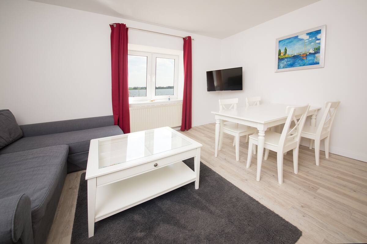 Una sala moderna con un sofá gris, una mesa de centro blanca y una mesa de comedor con sillas. Un televisor de pantalla plana está montado en la pared, y un cuadro se exhibe sobre el área del comedor.