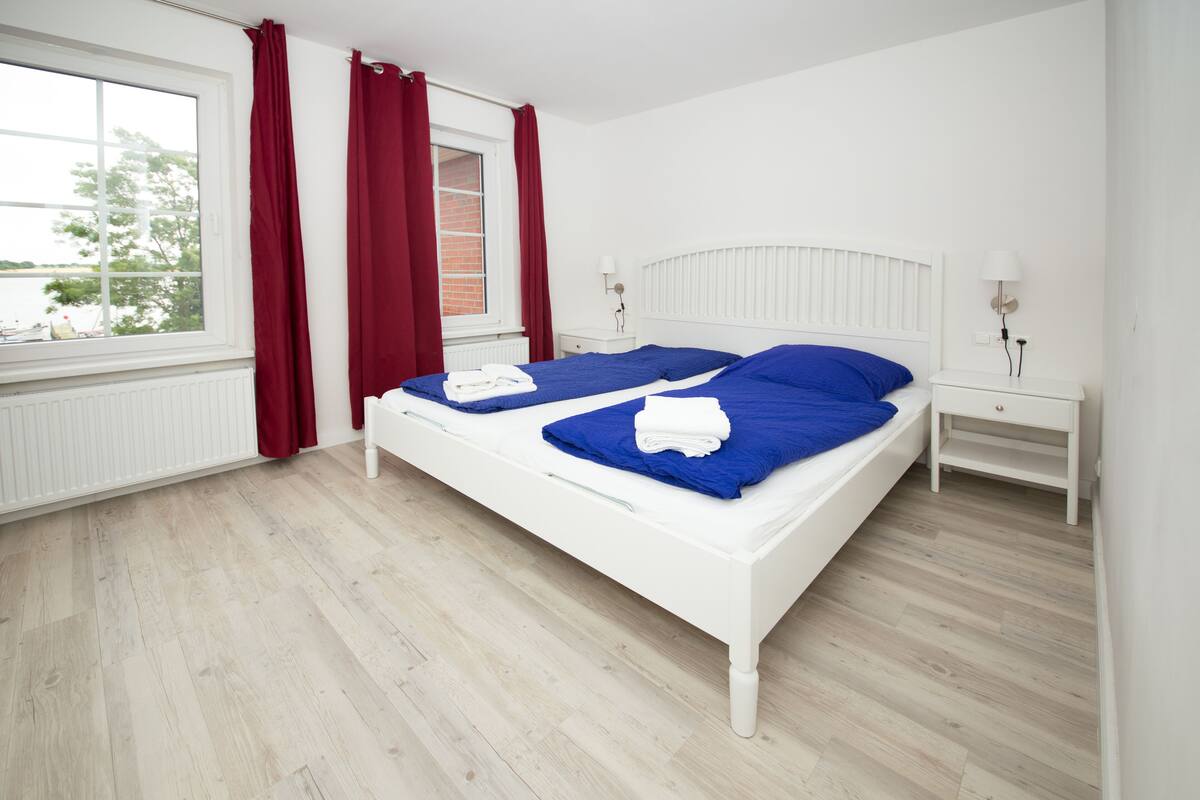 Cama doble con ropa de cama azul, cabecera blanca y mesita de noche con lámpara.