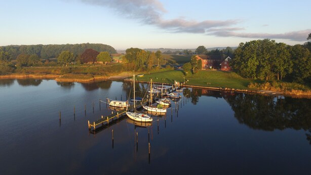 A calm lake with boats docked at a pier, surrounded by greenery and a few buildings.