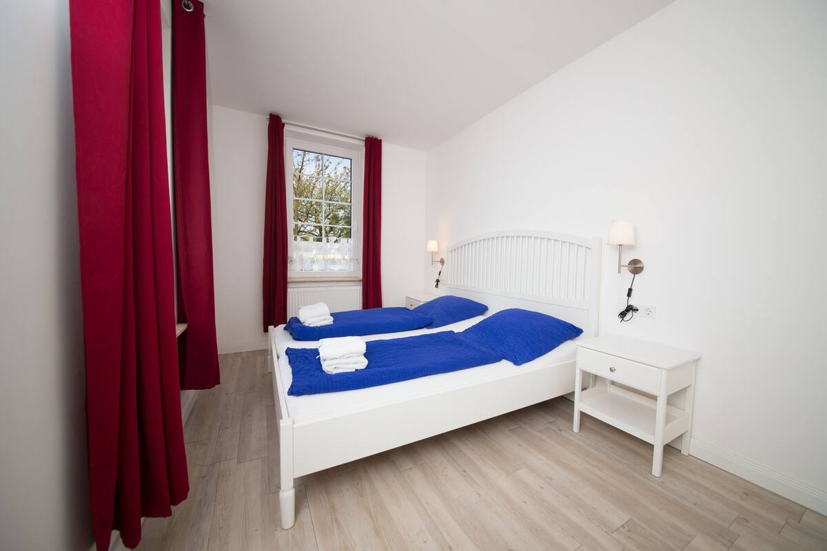 Una cama blanca con ropa de cama azul, una mesita de noche blanca y una ventana con cortinas rojas.