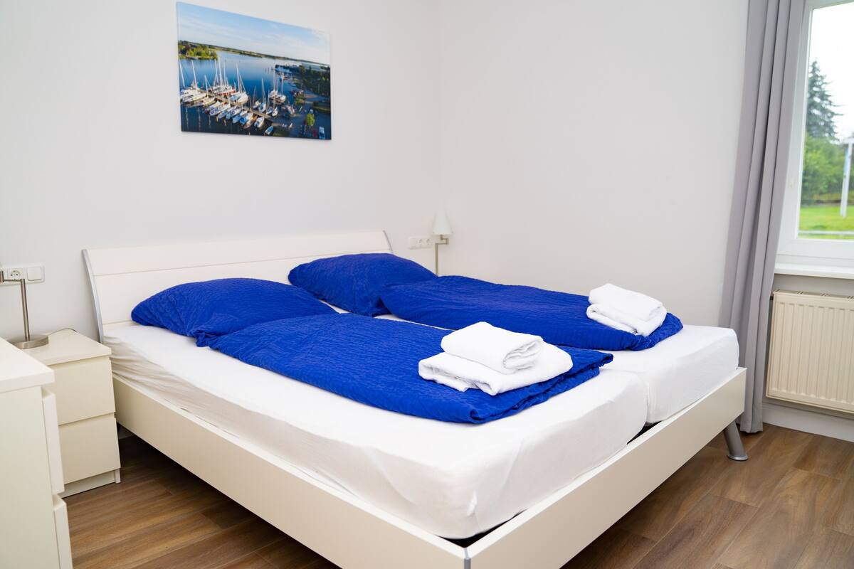 Cama doble con ropa de cama azul y sábanas blancas, una mesita de noche con lámpara y una ventana con vista a zonas verdes.