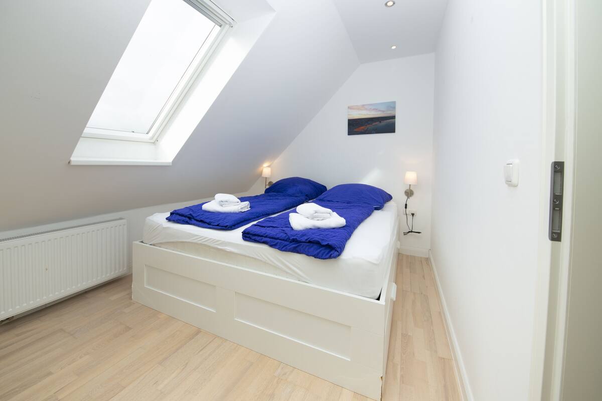 Un dormitorio con techo inclinado, tragaluz, cama blanca con ropa de cama azul, mesita de noche con lámpara y radiador.