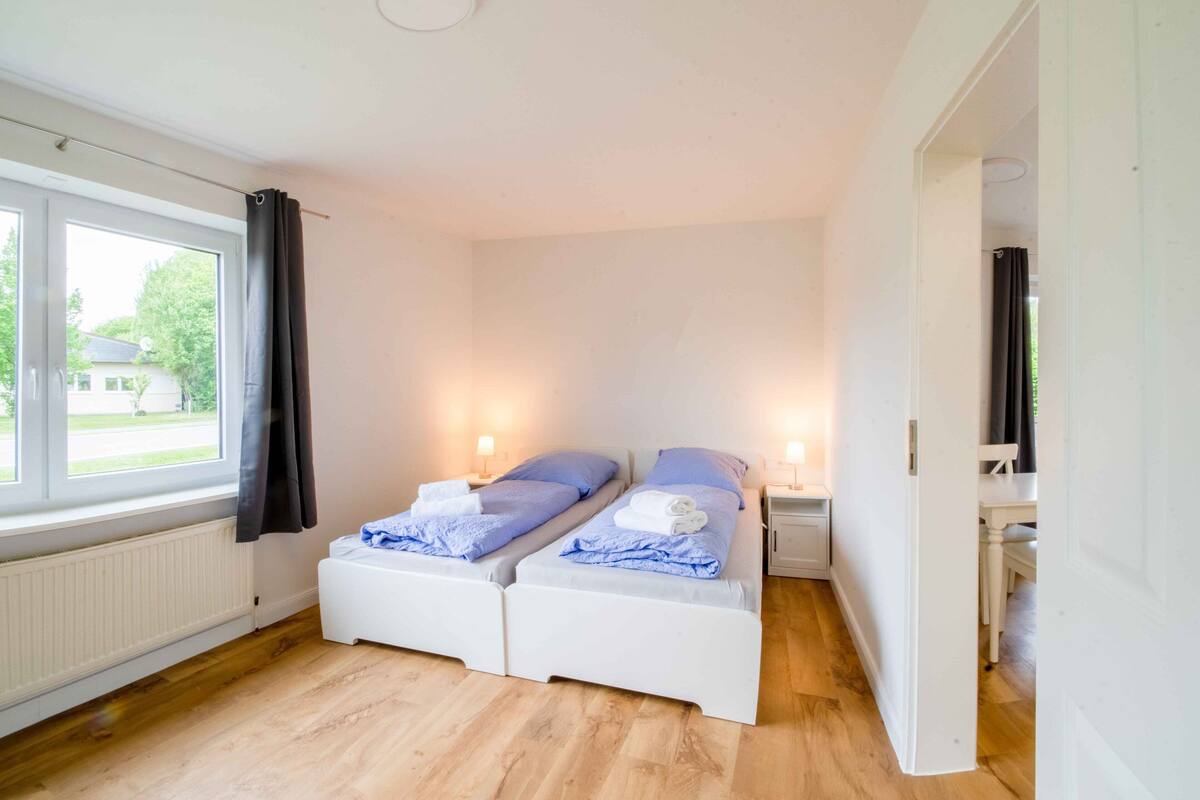 Una habitación pequeña y luminosa con dos camas individuales, una ventana con cortinas, un radiador y una puerta blanca.