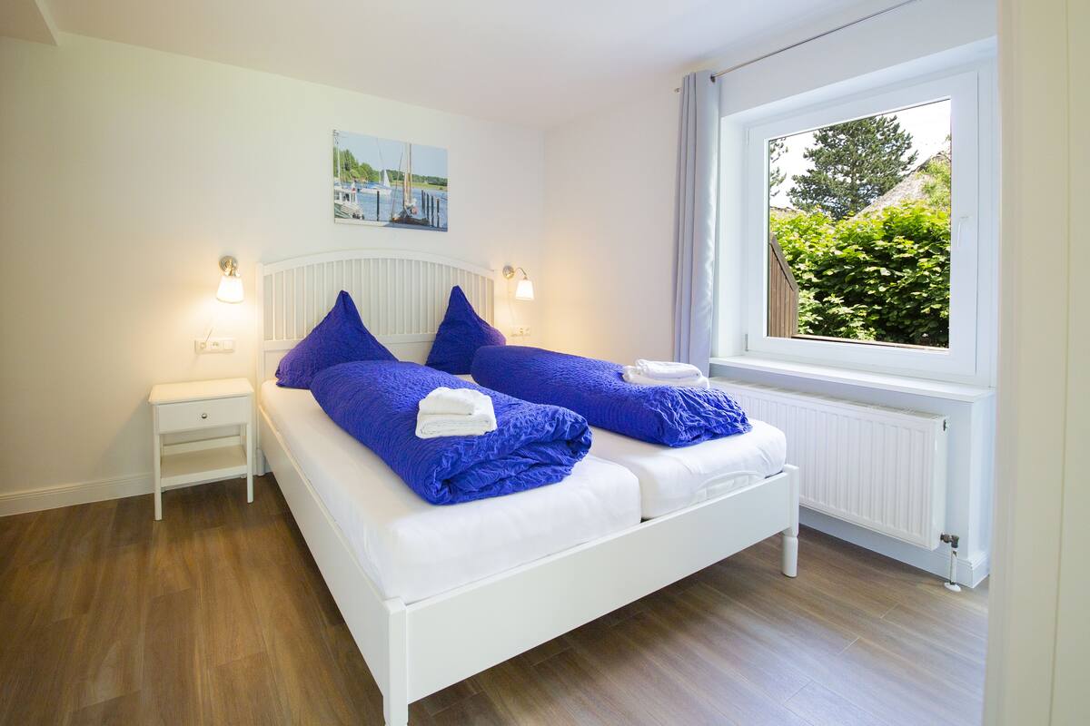 Un dormitorio con cama blanca, ropa de cama azul, una mesita de noche, una lámpara de pared y una ventana con vistas a la vegetación.