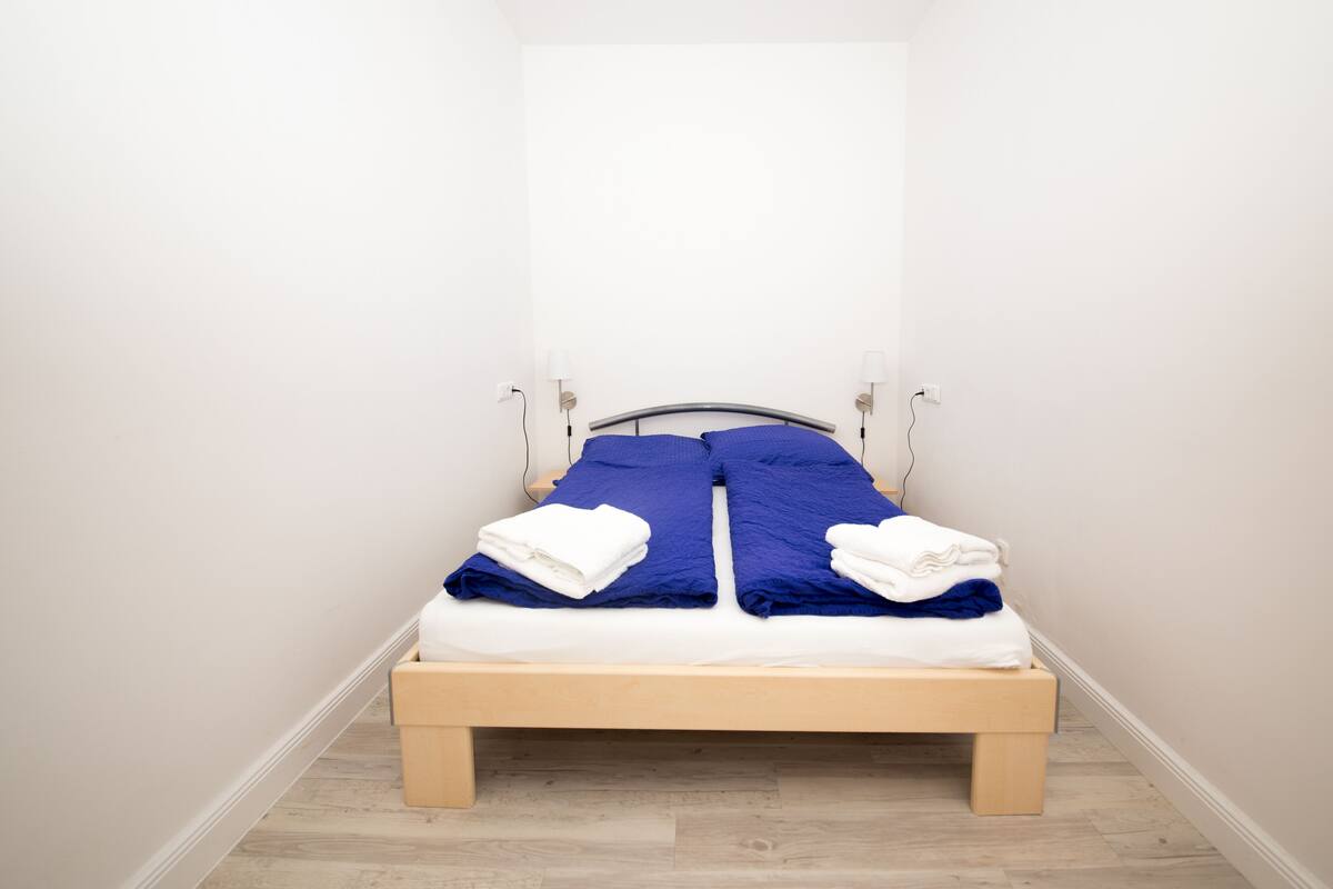 Una cama pequeña y bien hecha, con estructura de madera y ropa blanca, ubicada en una habitación con paredes blancas y piso de madera.
