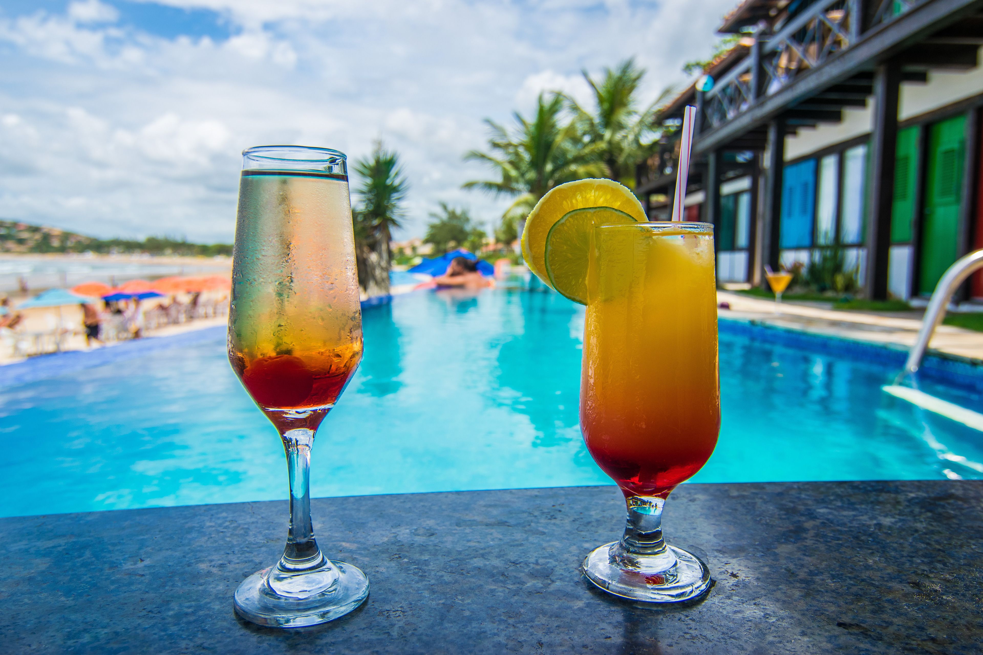 Two cocktails on a table with a pool and a beach in the background.