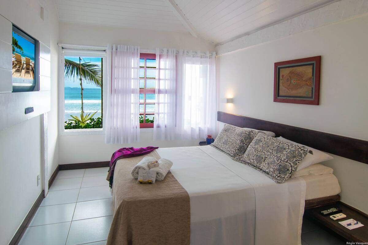 A hotel room with a bed, a window with curtains, a painting on the wall, and a view of the ocean.