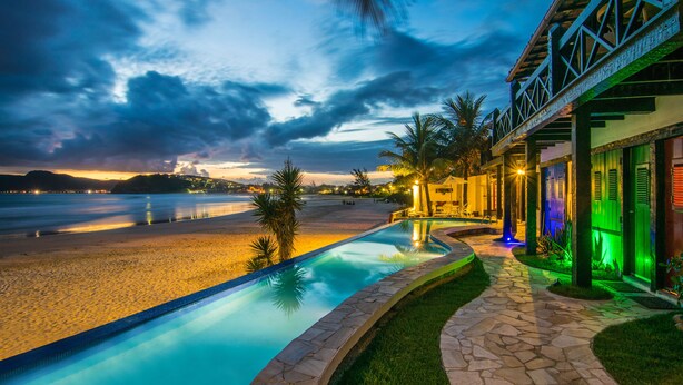 A pool area with a view of the beach and ocean, stone pathway, and a building with a balcony. - Chez Pitu Praia Hotel (Búzios)