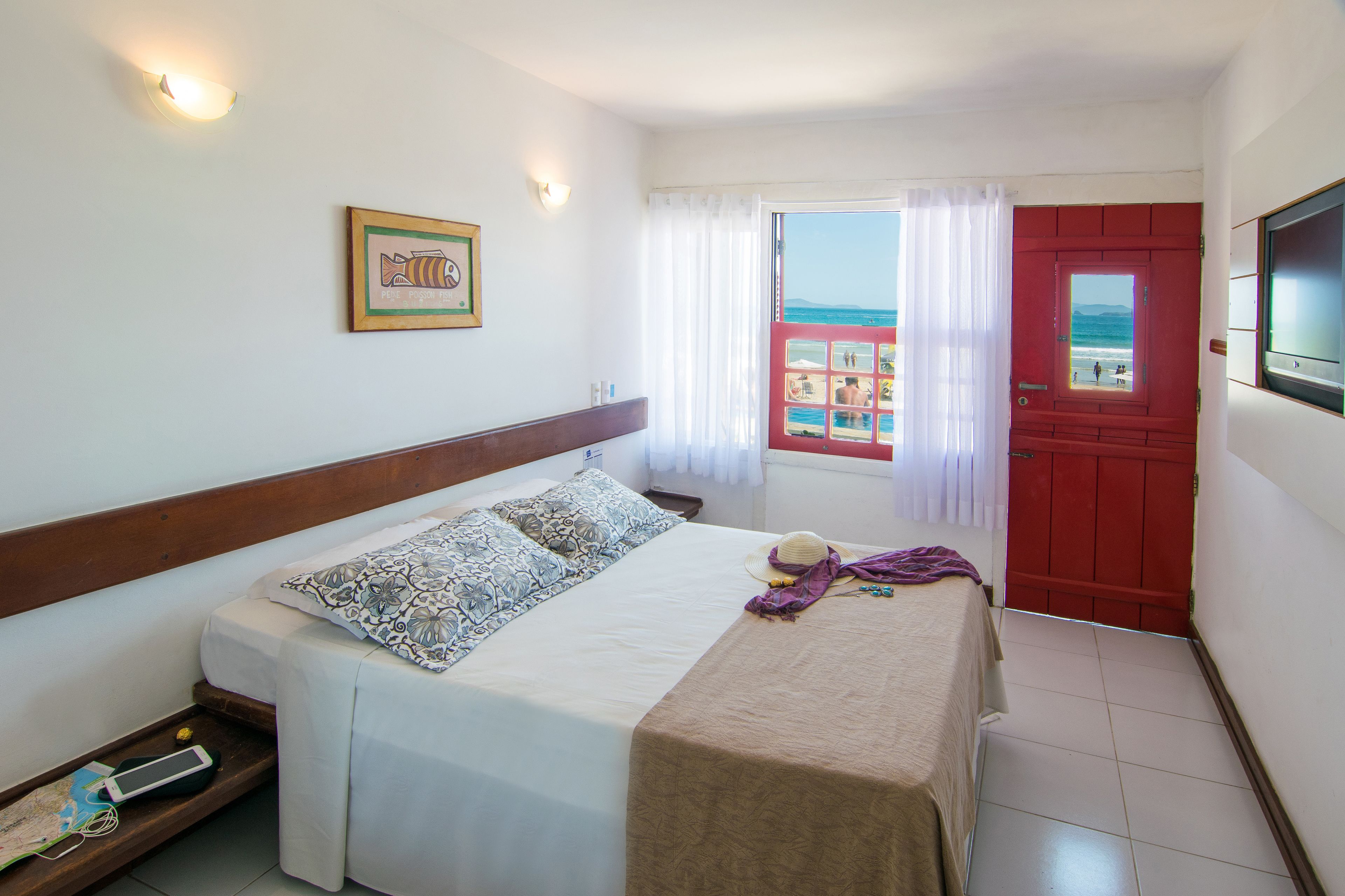 Une chambre à coucher avec un lit, une fenêtre donnant sur la plage, une porte rouge et un tableau encadré au mur.