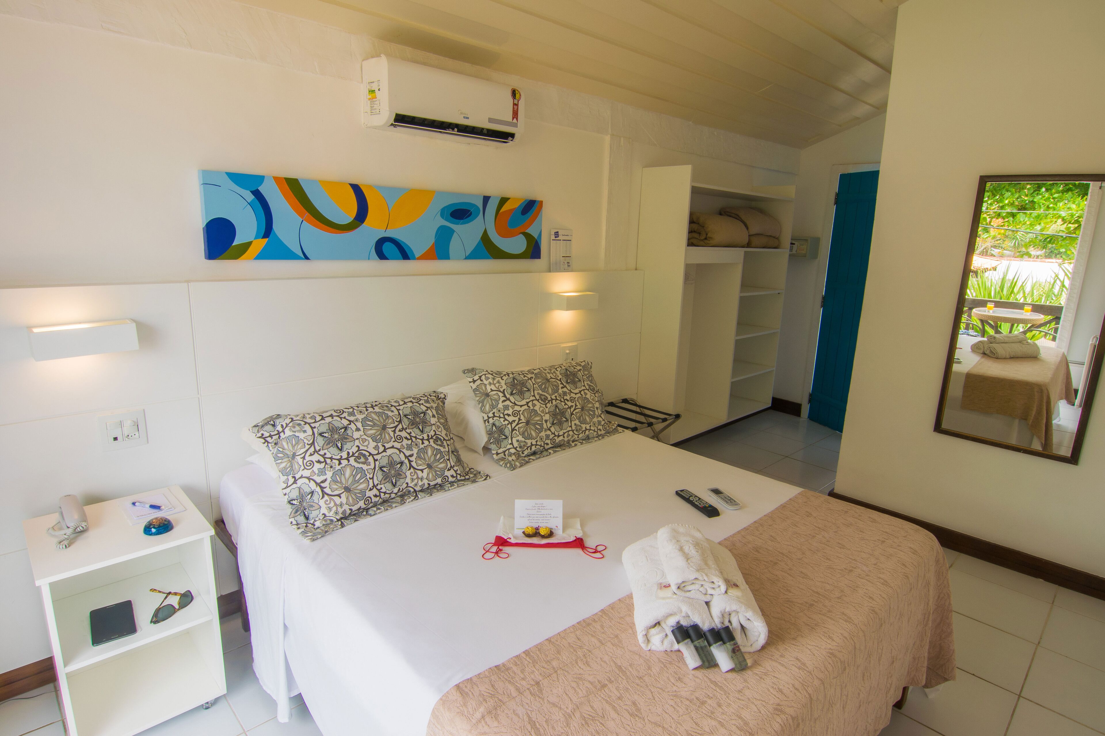 Ein Hotelzimmer mit Bett, Nachttisch, Wandairconditioner, einem farbenfrohen abstrakten Gemälde, einem Spiegel und Blick ins Freie.