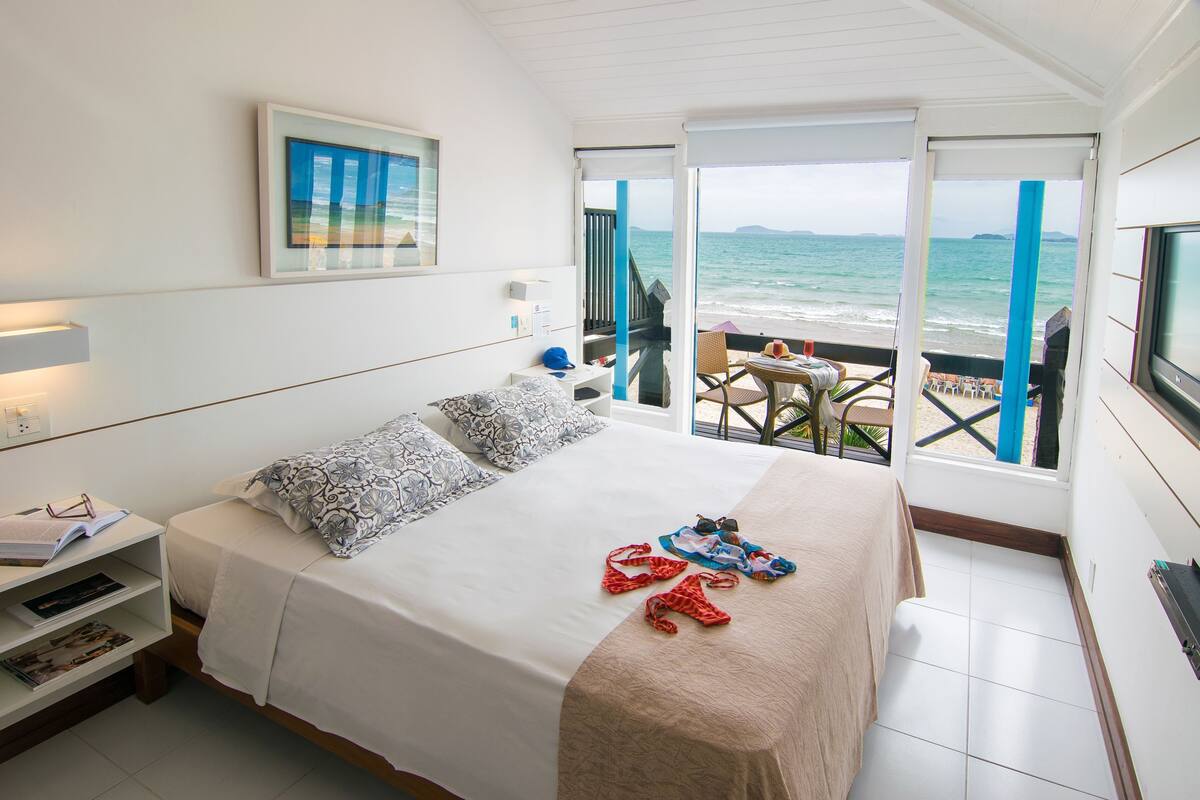 A bedroom with a bed, a nightstand, a window with a view of the sea, and a balcony with a table and chairs.
