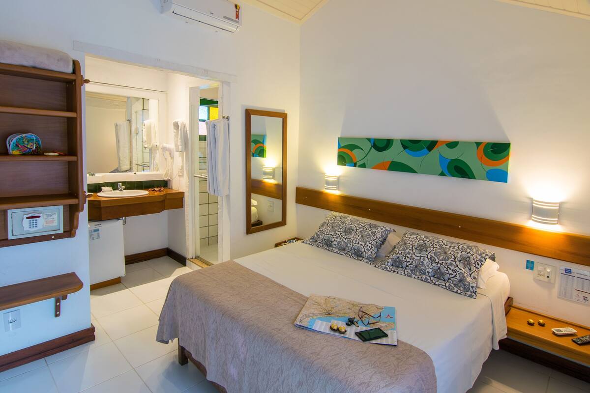 Une chambre d’hôtel avec un lit, une table de chevet, un miroir, une œuvre d’art accrochée au mur et une salle de bain visible à travers une porte ouverte.