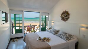 A bedroom with a bed, a table with a chair, and a view of the beach through open doors. - Chez Pitu Praia Hotel (Búzios)