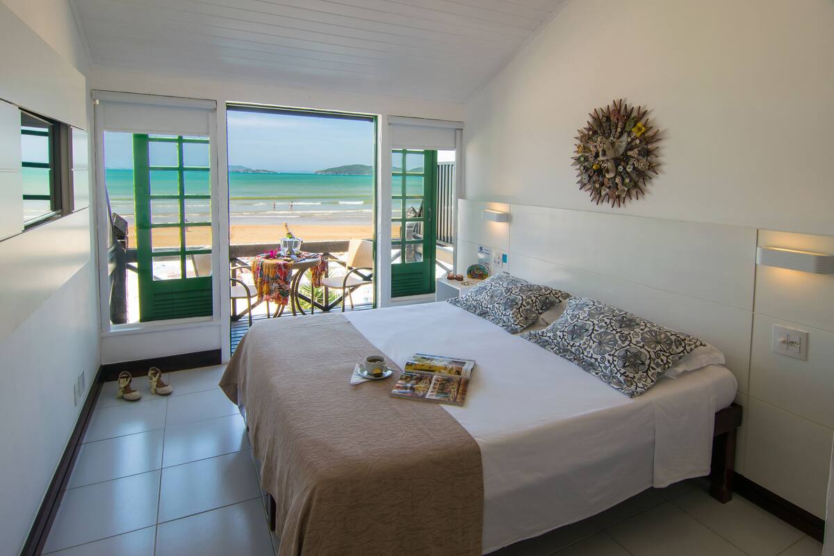 A bedroom with a bed, a table with a chair, and a view of the beach through open doors.