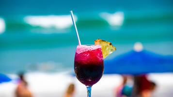 Um cocktail com canudo e uma fatia de abacaxi na borda, com uma praia ao fundo, incluindo pessoas e guarda-sóis.