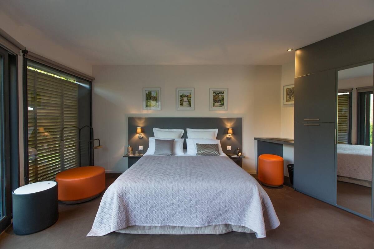 A modern bedroom with a large bed, two bedside tables, a nightstand, and a seating area with a round ottoman.