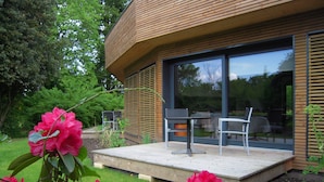 Una casa moderna con una terraza de madera, un área de comedor con dos sillas y un ventanal grande.