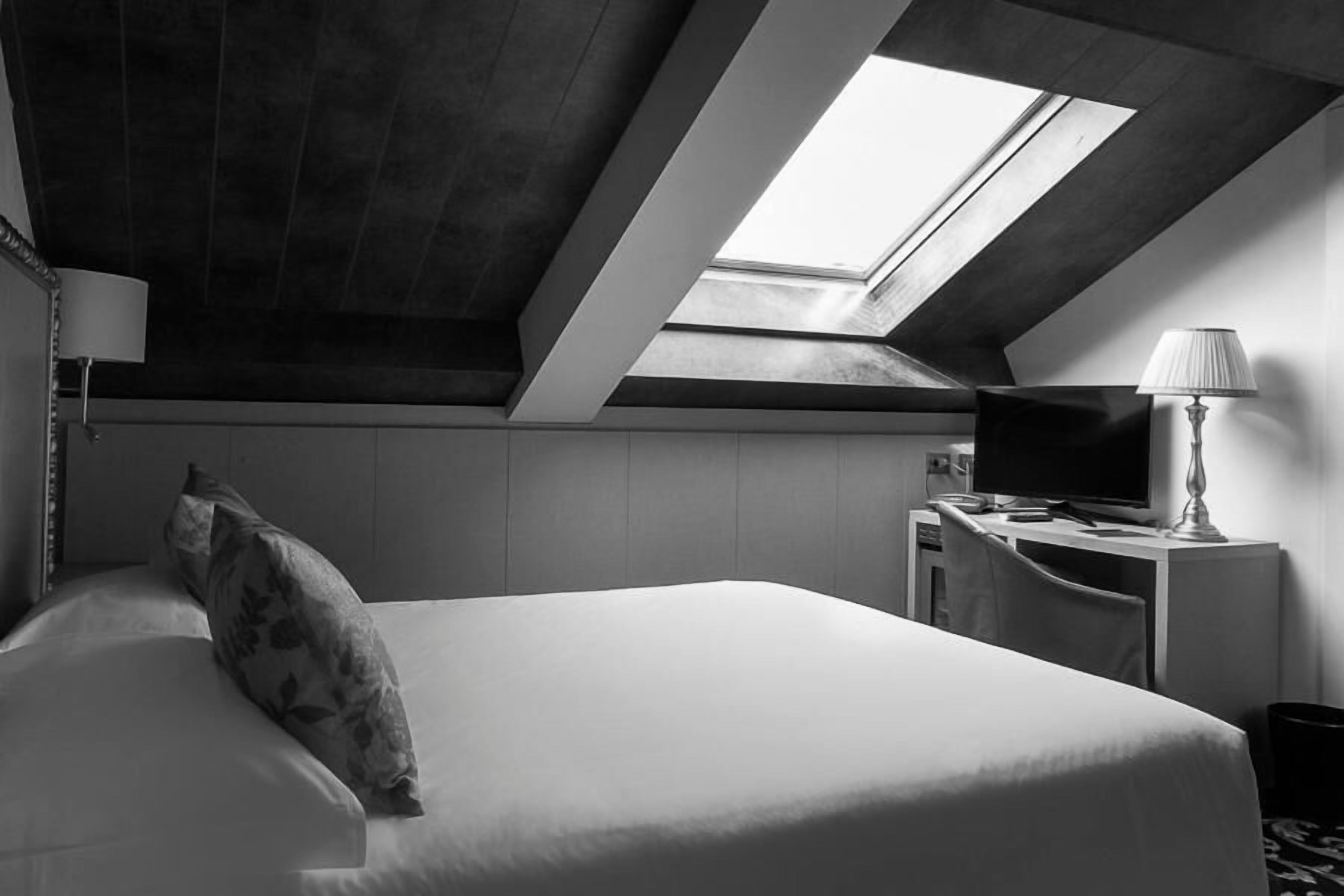 A modern hotel room with a skylight, a bed with a floral pillow, a desk with a lamp, and a television.