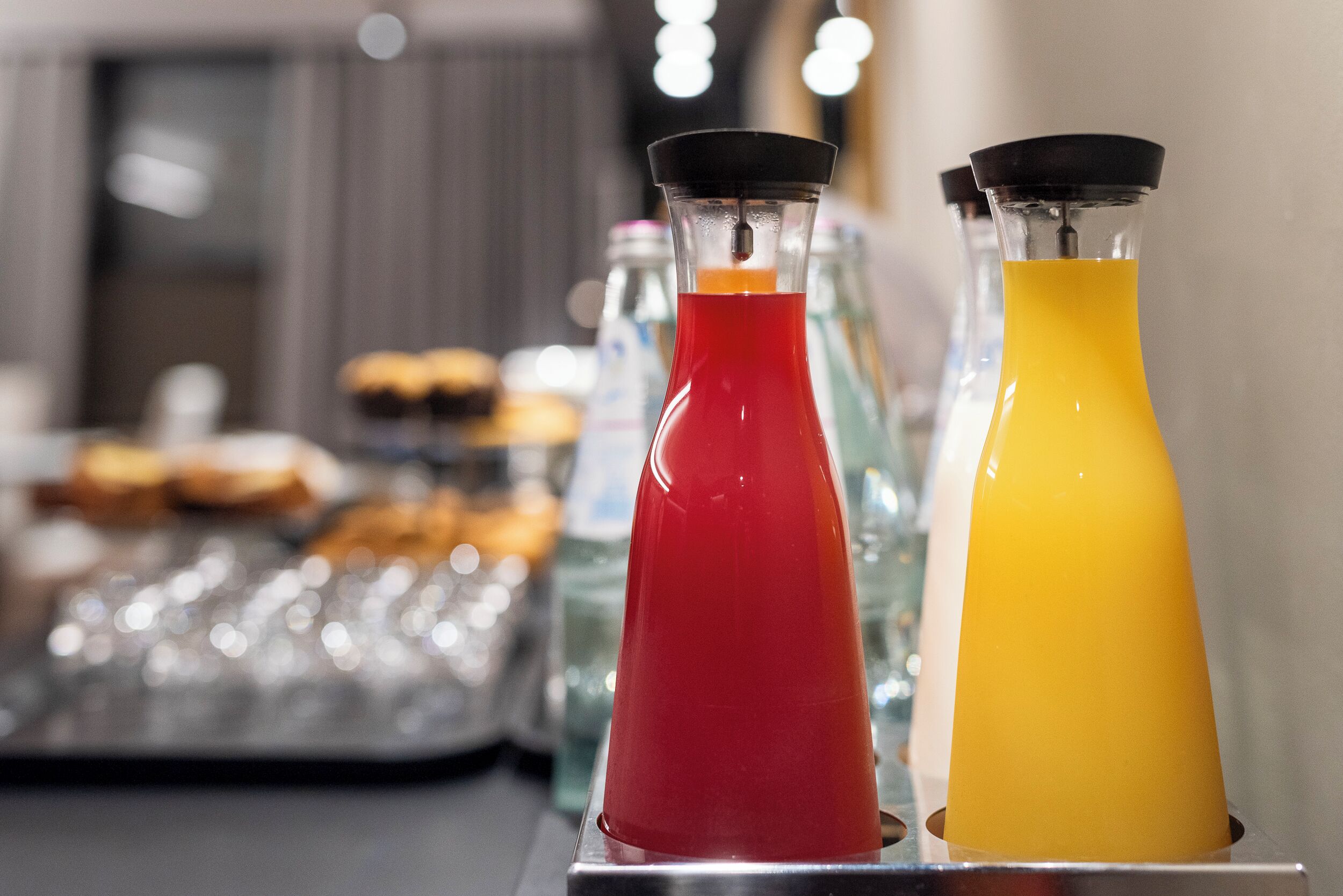 Deux distributeurs de boissons contenant des liquides rouges et jaunes, un plateau de pâtisseries et un arrière-plan flou d’une pièce avec une fenêtre.