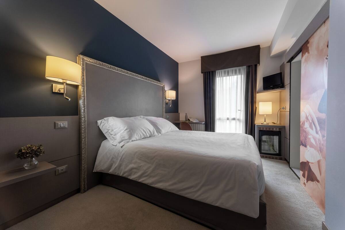 Habitación de hotel con una cama grande, dos mesitas de noche, un televisor de pantalla plana, un escritorio con lámpara y un ventanal con cortinas.