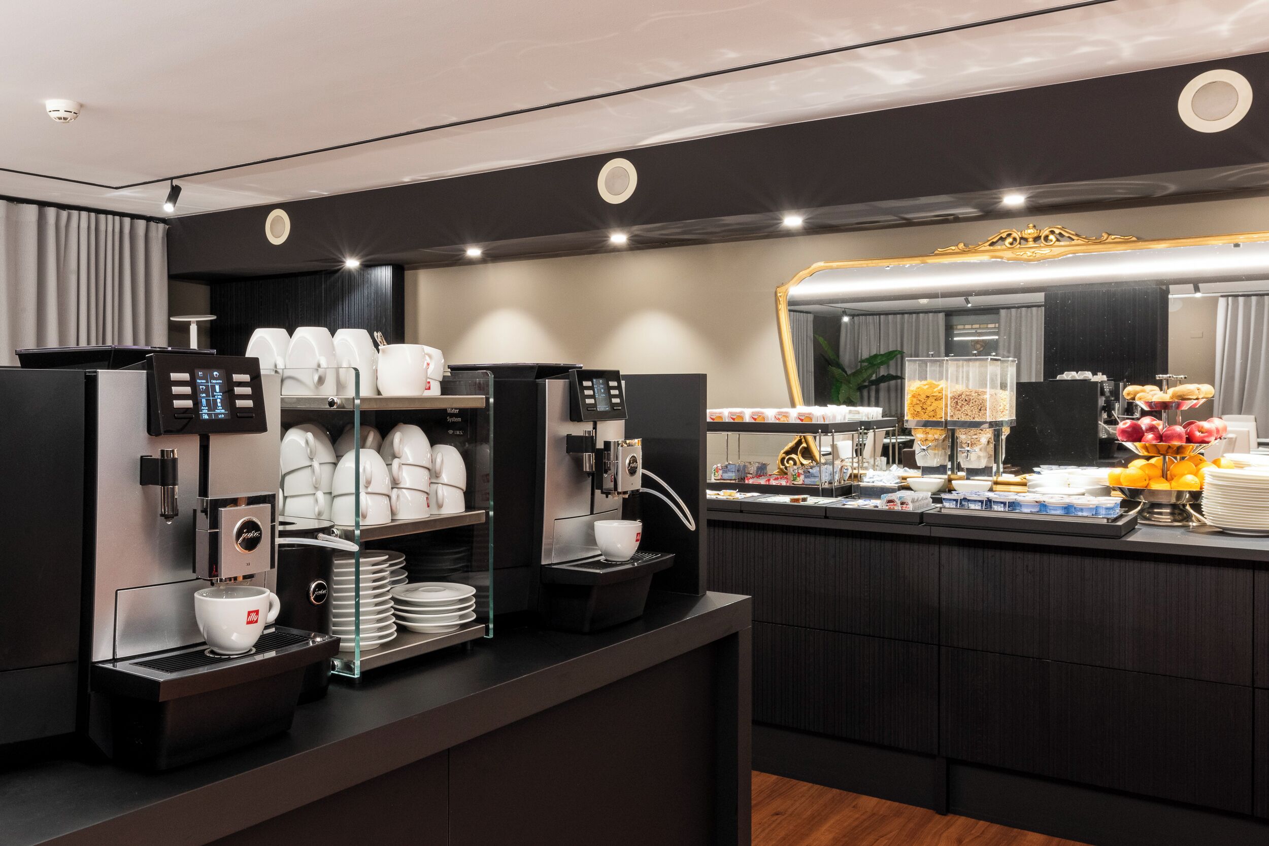 A modern coffee station with multiple espresso machines, a variety of coffee cups, and a buffet with fruits and pastries.
