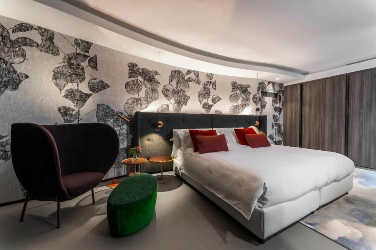 Un dormitorio moderno con una pared curva, una cama grande con almohadas rojas, un sillón oscuro y un reposapiés verde.