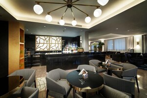 Un moderno vestíbulo de hotel con una zona de bar, asientos y una lámpara decorativa en el techo.