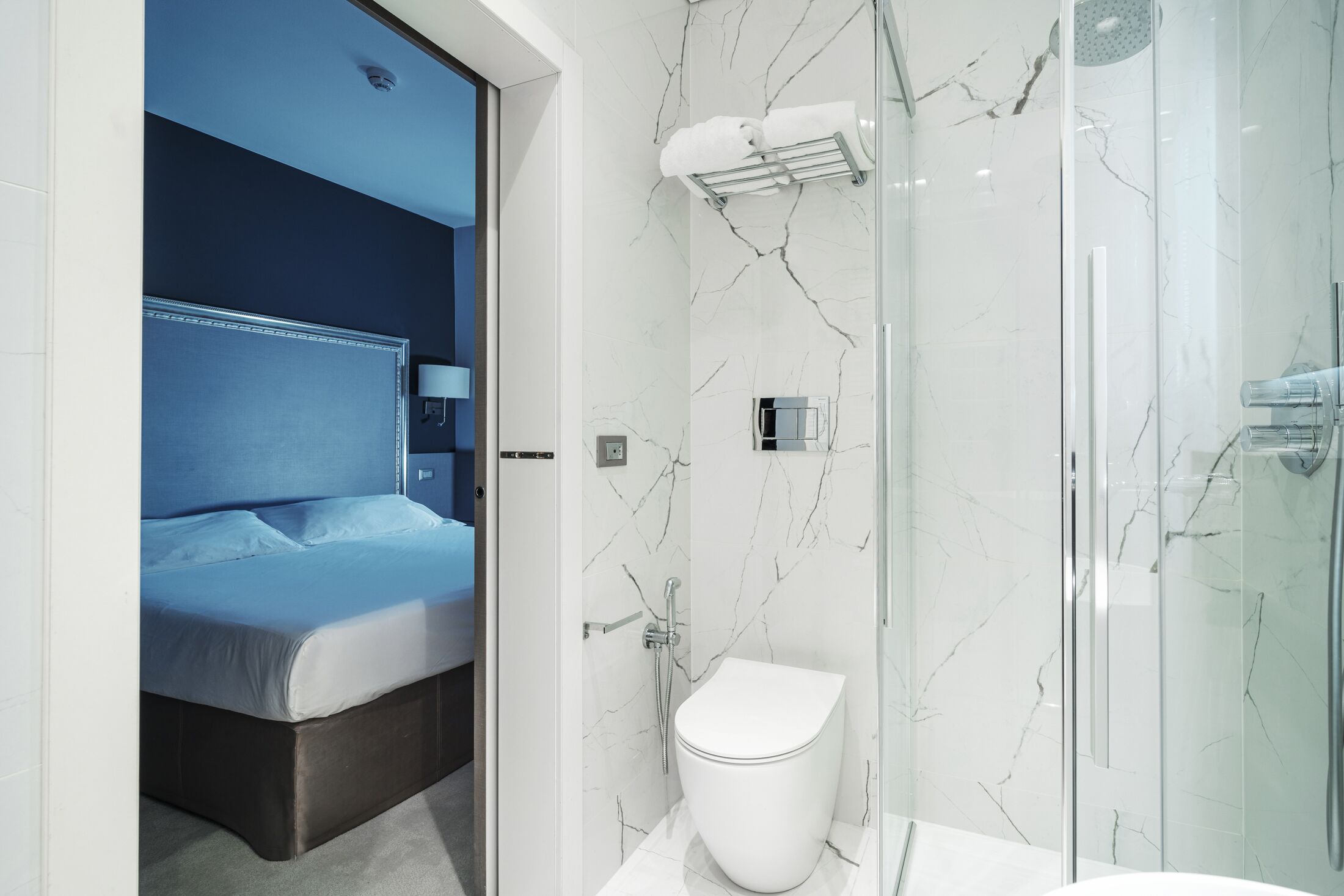 Un bany modern amb una cabina de dutxa de vidre, un vàter blanc i un llit amb el capçal blau.